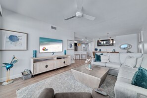 Ein modernes Wohnzimmer mit einem weißen Sofa, einem Couchtisch und einem Fernseher. Es gibt einen Deckenventilator, eine Holzkommode und eine dekorative Vogel-Skulptur.