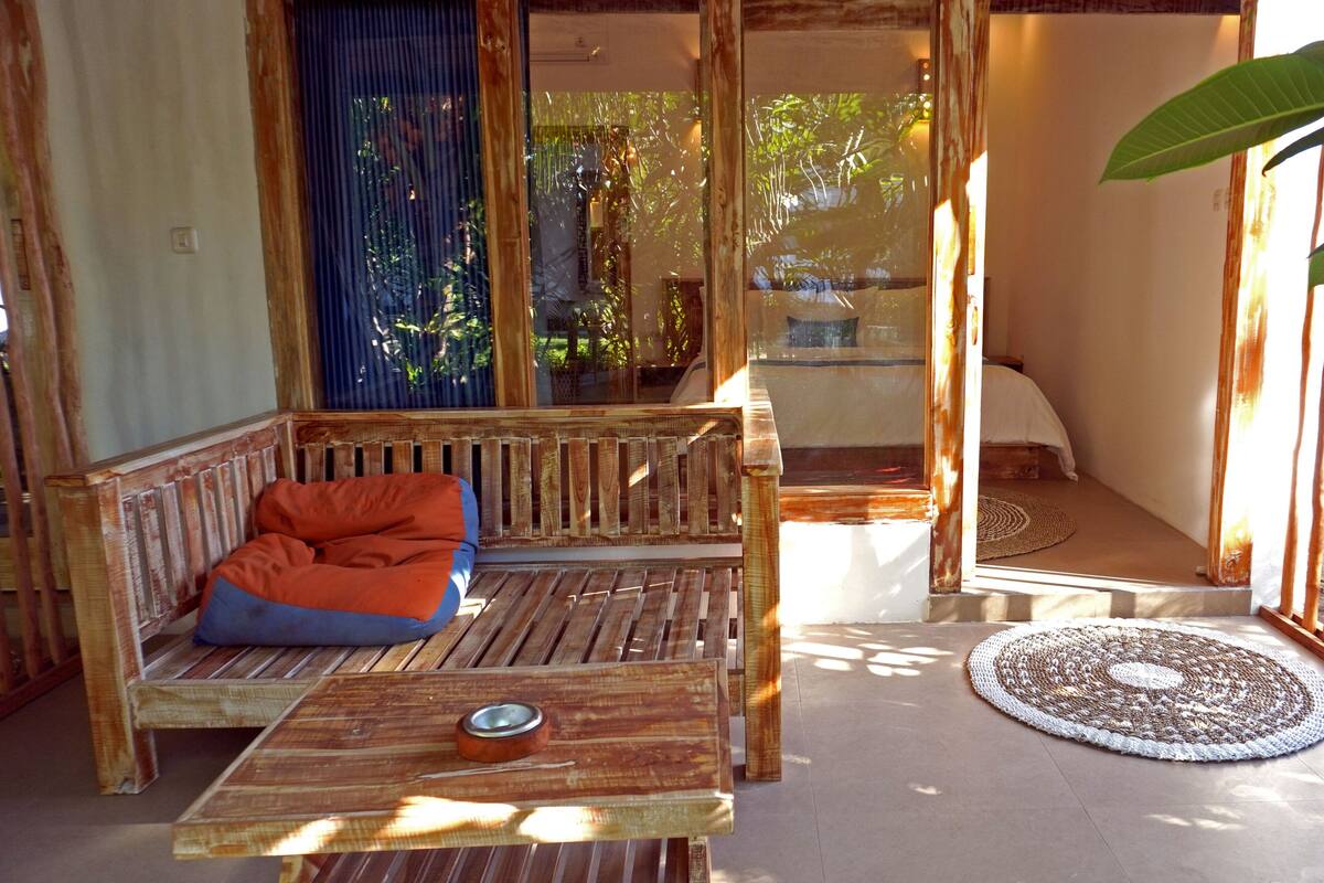 Un espace extérieur aménagé avec des meubles en bois, comprenant un banc et une table, un tapis tissé, et une vue sur la verdure par une fenêtre.