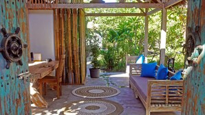 A covered outdoor seating area with a wooden table, chairs, and a sofa with blue cushions. There are woven mats on the floor and a potted plant in the background.