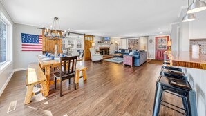 Amplia sala de estar con mesa de comedor, sillas de madera, sofá y una cocina con mostrador de madera y taburetes de bar.
