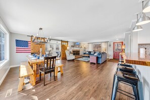 A spacious living area with a dining table, wooden chairs, a sofa, and a kitchen area with a wooden countertop and bar stools. - Lake Point Retreat - Lakefront Home with Hot Tub, Fire pit & Game room (Huddleston)