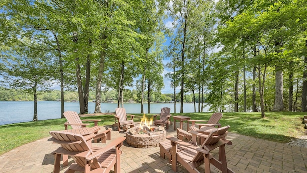 Una fogata rodeada de sillas Adirondack, ubicada en un patio de ladrillo con vista al lago.