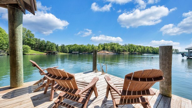 Un muelle de madera con dos sillas Adirondack mirando a un lago, rodeado de árboles y un cielo despejado.
