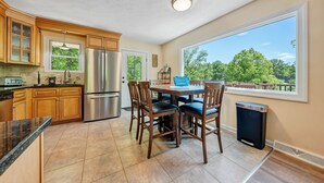 Una cocina con armarios de madera, un refrigerador de acero inoxidable, una mesa de comedor con sillas y un ventanal con vistas a árboles.