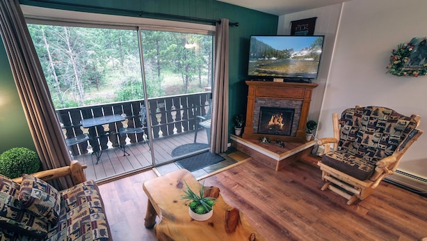 A cozy living room with a fireplace, a television, and a view of a forest through the sliding glass door. - One Bedroom Condo only Steps from Fall River (Estes Park)