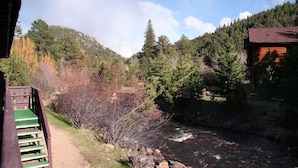 A cabin with a wooden exterior, a stone pathway leading to a wooden deck, and a river flowing through a forested area. - One Bedroom Condo only Steps from Fall River (Estes Park)