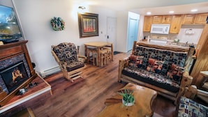 A cozy living room with a fireplace, wooden furniture, and a kitchen area. - One Bedroom Condo only Steps from Fall River (Estes Park)