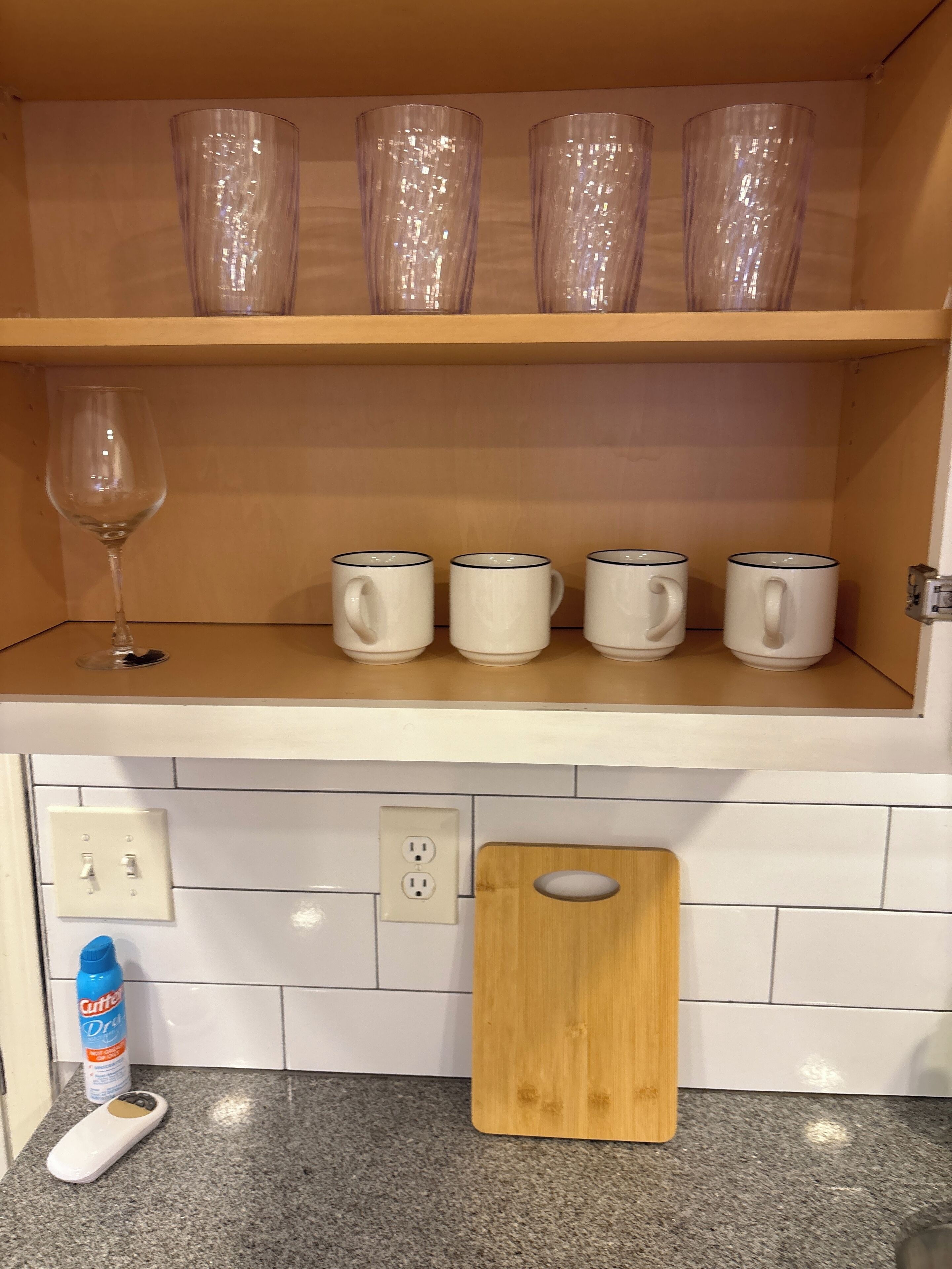 A kitchen with a countertop, a wooden cutting board, a wine glass, and four white mugs on a shelf.