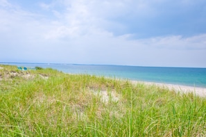 A beach with sand and grass in the foreground, a clear sky, and a calm sea in the background.