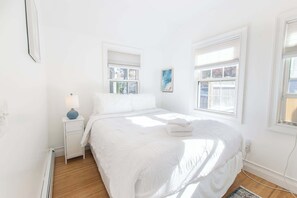 Une chambre à coucher blanche, avec un lit, une table de chevet, une lampe et deux fenêtres.