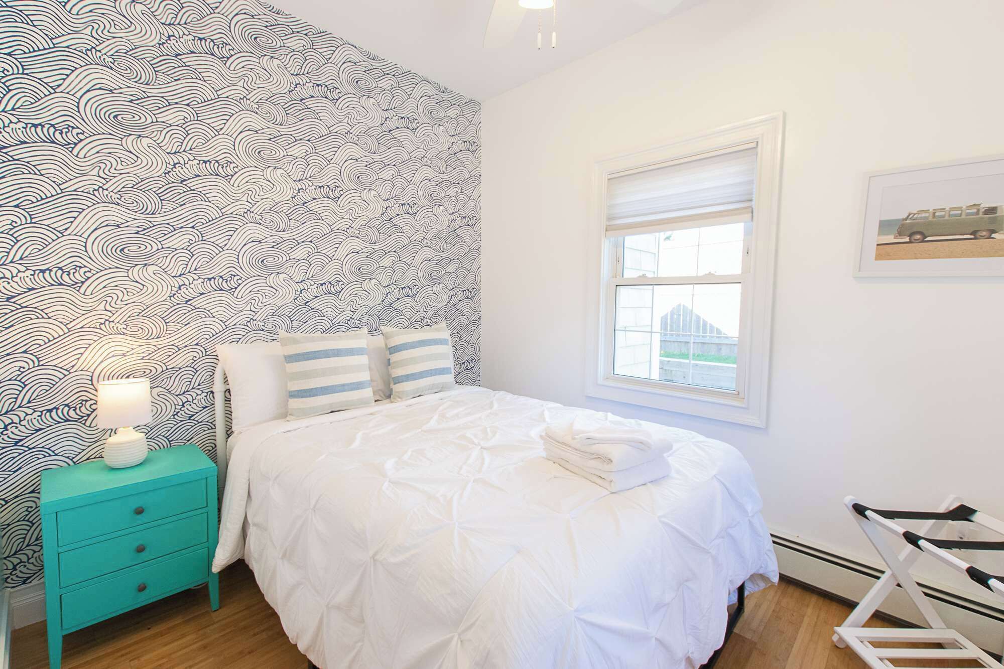 Un dormitorio con papel tapiz estampado, una cama con ropa blanca, una mesita de noche turquesa con una lámpara y una ventana con vistas.