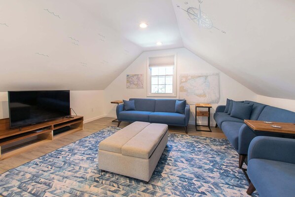 Un salon chaleureux aménagé dans les combles, avec un téléviseur, des canapés bleus, un meuble TV en bois et un tapis à motifs.