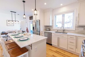 A modern kitchen with white cabinets, a central island, stainless steel appliances, and pendant lighting. - Walk to Water with Hot Tub! (Yarmouth)