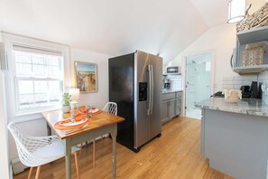 A compact kitchen with a stainless steel refrigerator, a dining table with orange placemats, and a white chair. - Walk to Water with Hot Tub! (Yarmouth)