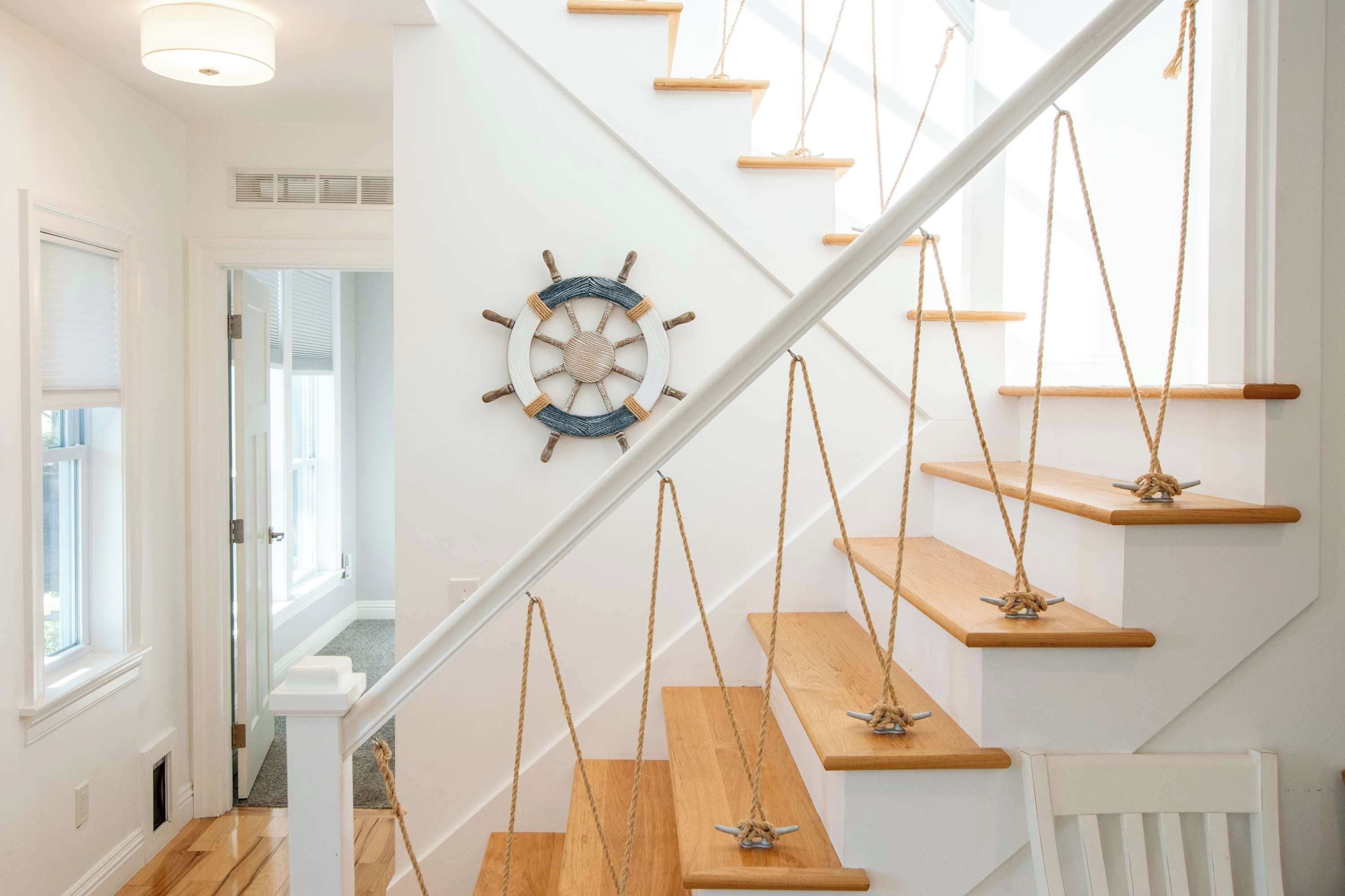 A staircase with wooden steps and rope swings, a white wall with a ship's wheel decoration, and a door leading to another room.