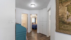 Un pasillo con un aparador verde azulado, paredes blancas y una lámpara en el techo. Se ve una puerta que da a una habitación con una cama y una ventana con cortinas azules.