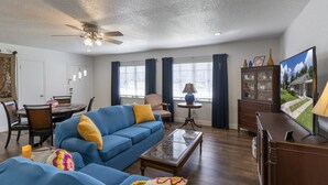 Una sala de estar con un sofá azul, una mesa de centro, un televisor, un aparador y un comedor con una mesa y sillas.