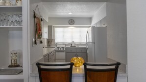 Cocina pequeña con armarios blancos, estufa, lavabo y ventana con persianas. Hay dos sillas en la mesa del comedor.