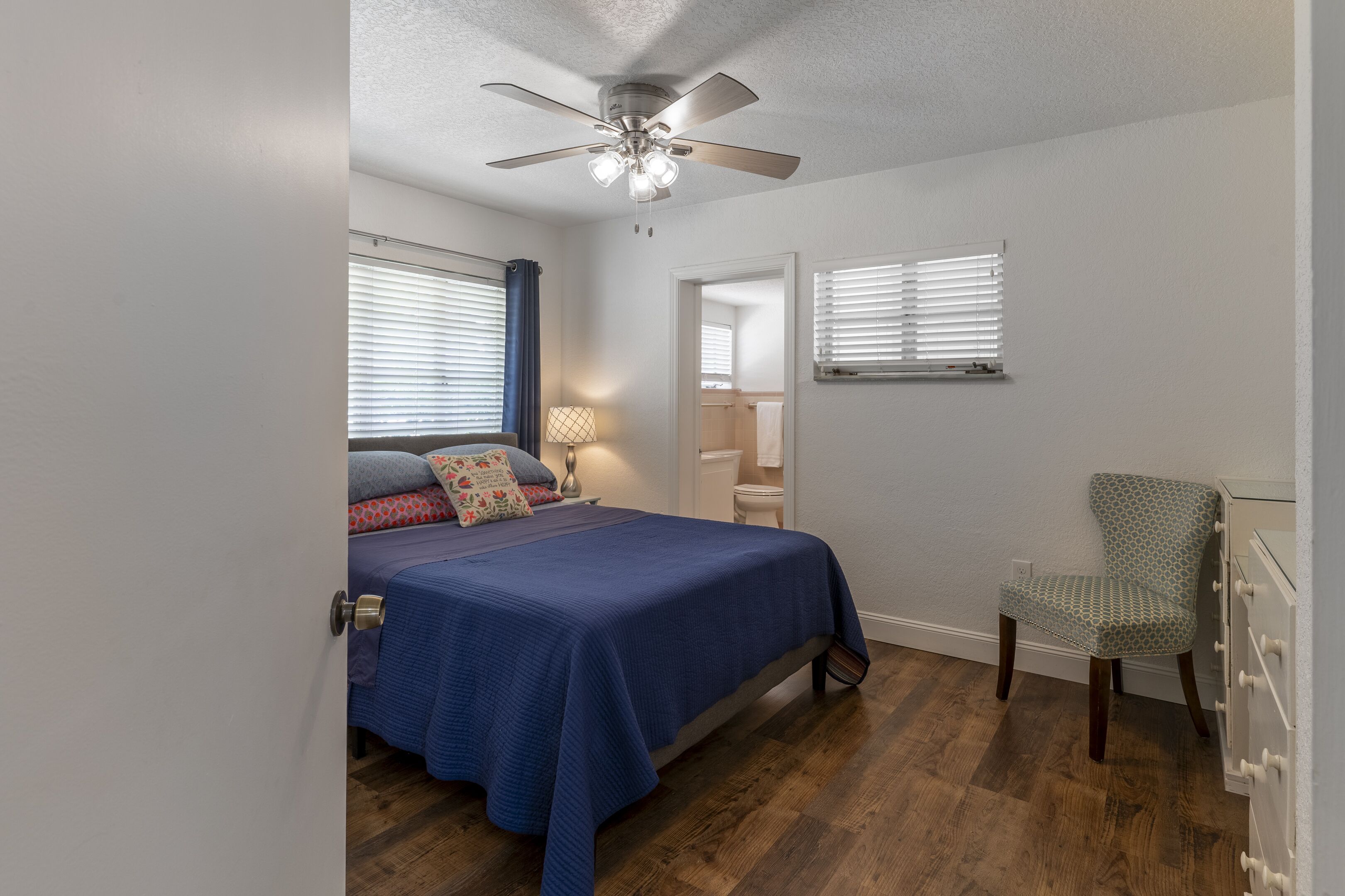 Une chambre à coucher comprenant un lit, une chaise, un ventilateur de plafond et une fenêtre avec des stores.