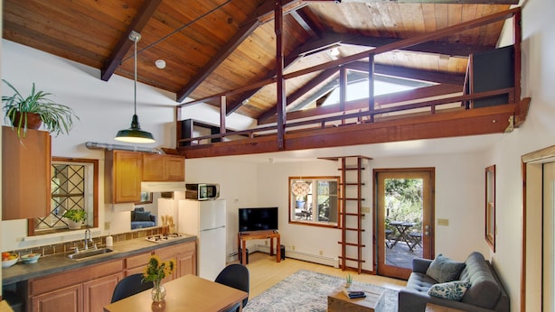 Um amplo loft com pé direito alto, vigas de madeira, cozinha com pia e micro-ondas, área de jantar com mesa e cadeiras, sala de estar com sofá e uma varanda com vista.