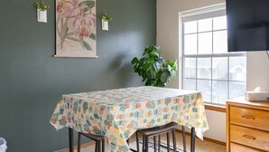 Un comedor con una mesa cubierta con un estampado de piña, una planta grande en maceta, una obra de arte floral enmarco y un televisor sobre un pedestal.