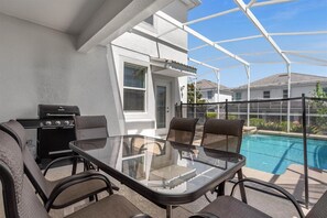 Une terrasse couverte avec une table et des chaises en verre, un barbecue et une piscine en arrière-plan.