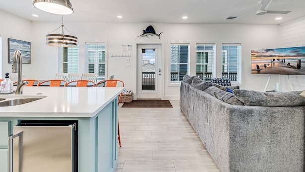 A modern kitchen with a central island, a dining area with orange chairs, and a large gray sofa.