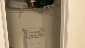 Ein kleiner Schrank mit weißer Tür, ein grüner Teppich, eine weiße Kommode mit Schubladen, ein weißer Korb und ein Regal mit gefalteter Kleidung und einer Kiste.