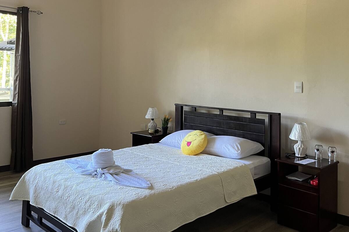 Ein ordentlich bezogenes Bett mit weißer Decke und Kissen, ein Nachttisch mit Lampe sowie ein Fenster mit Vorhängen.