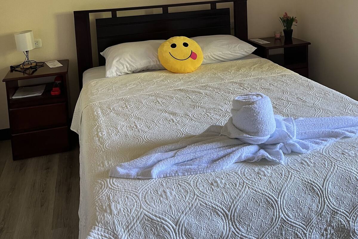Ein ordentlich bezogenes Bett mit einem gemusterten Überwurf, einem Kissen mit einem Smiley, einem weißen Handtuch und einem kleinen Tisch mit einer Lampe und einer Blumenvase.