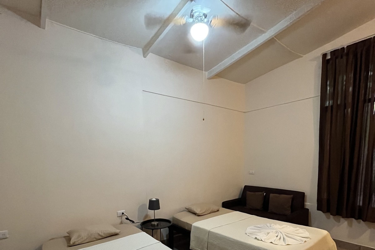 Ein Hotelzimmer mit zwei Einzelbetten, einem Deckenventilator, einem kleinen Tisch mit Lampe und einem Fenster mit Vorhängen.