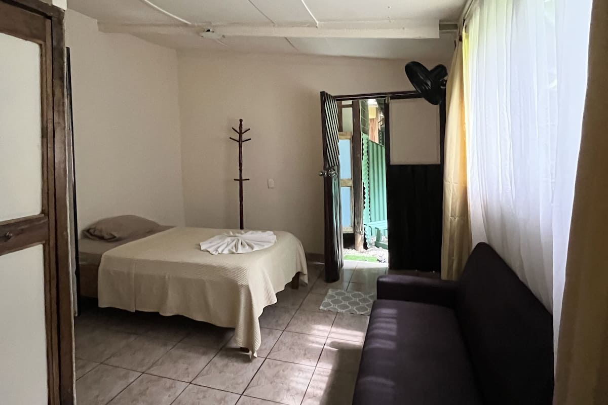 Ein Zimmer mit einem Bett, einem Sofa, einem Kleiderständer und Blick auf einen Garten durch ein Fenster.