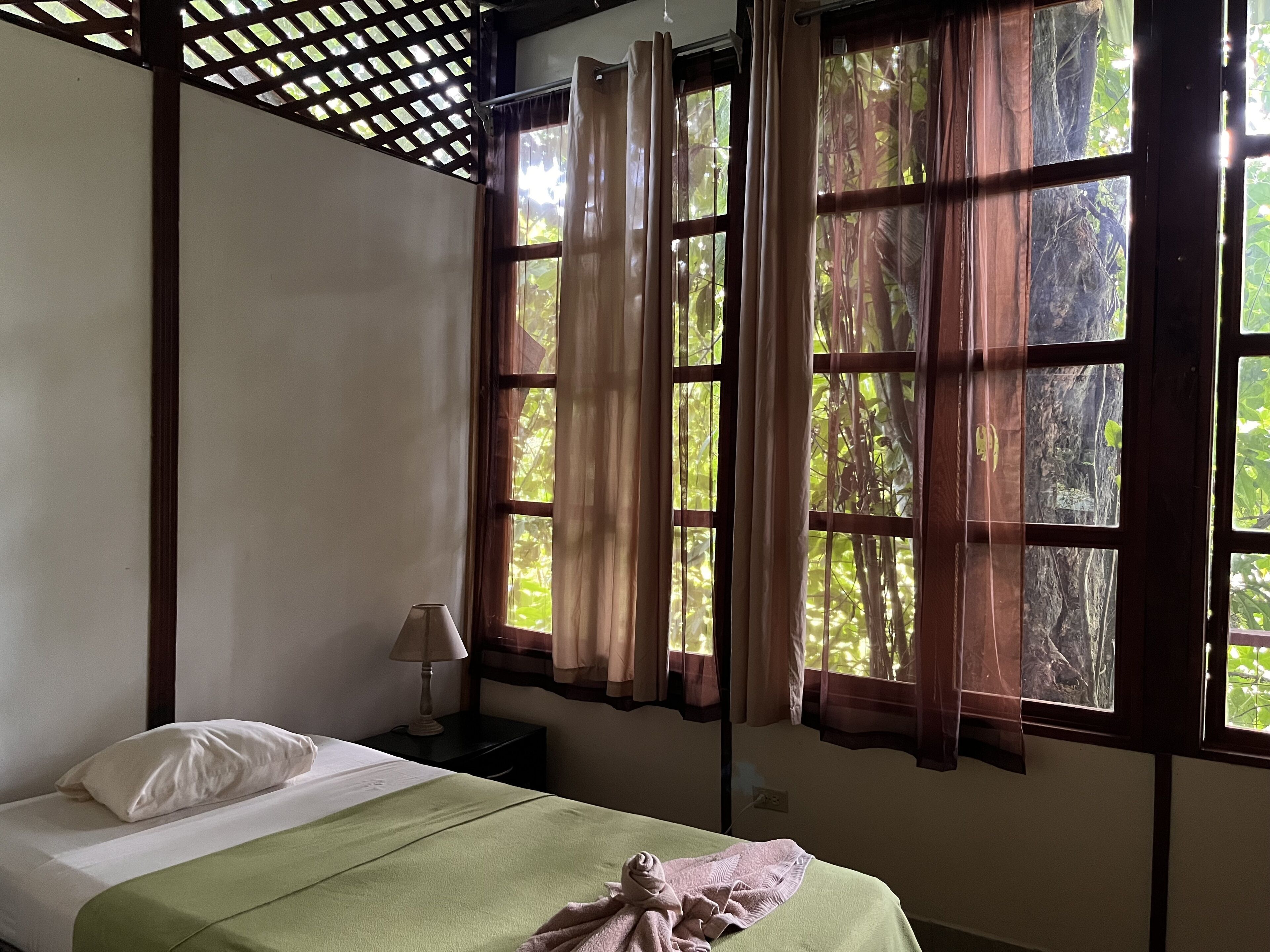 A room with a bed, a nightstand, a lamp, and large windows with curtains.