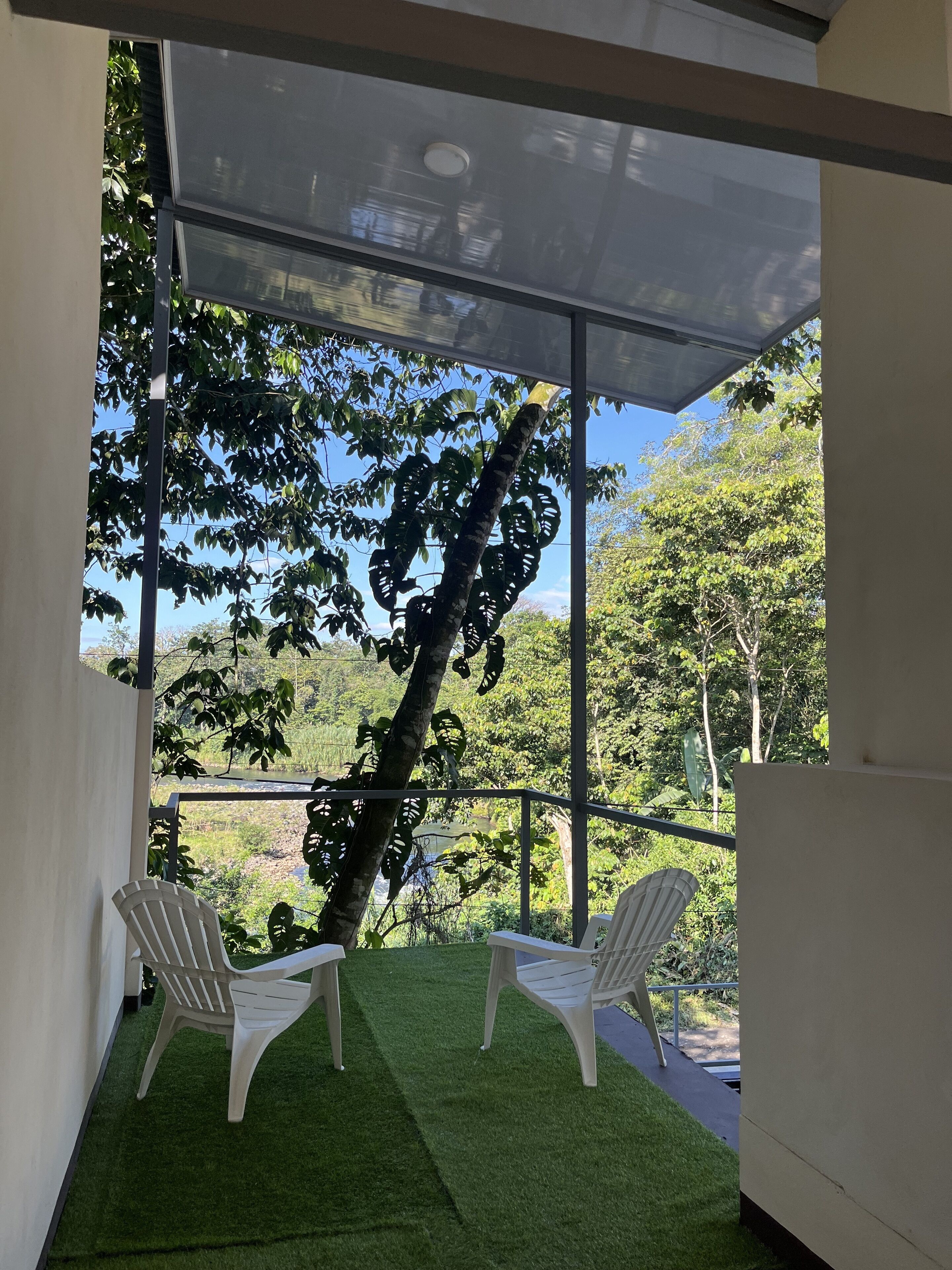 Un área de estar al aire libre techada con dos sillas de plástico blanco y una pared de vidrio, con vista a un paisaje verde exuberante y árboles.