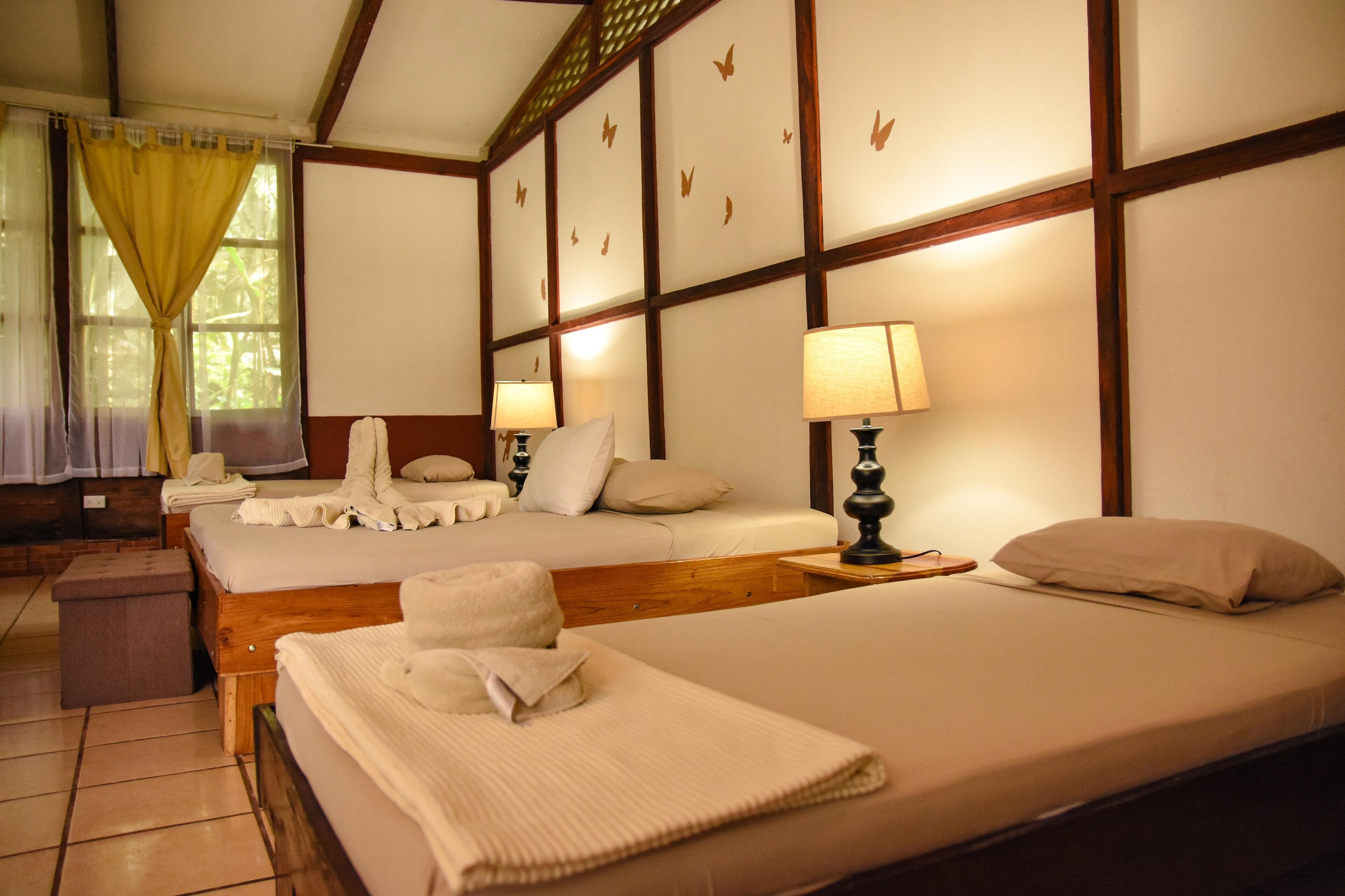 A hotel room with three beds, each with a pillow and a towel. There are two bedside lamps and a small wooden table.