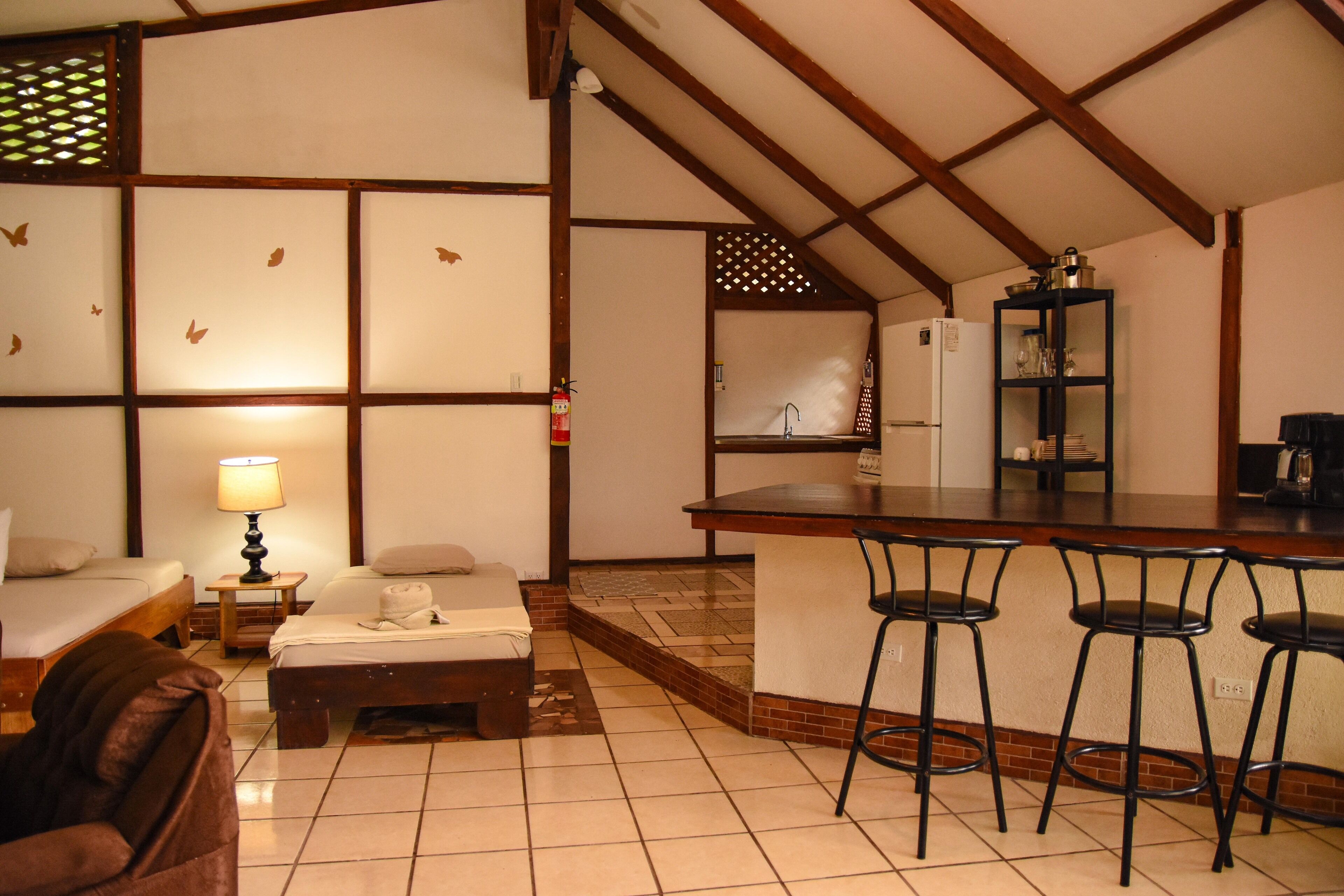 A hotel room with a kitchenette, a dining area with bar stools, a bed, a small table with a lamp, and a sofa.