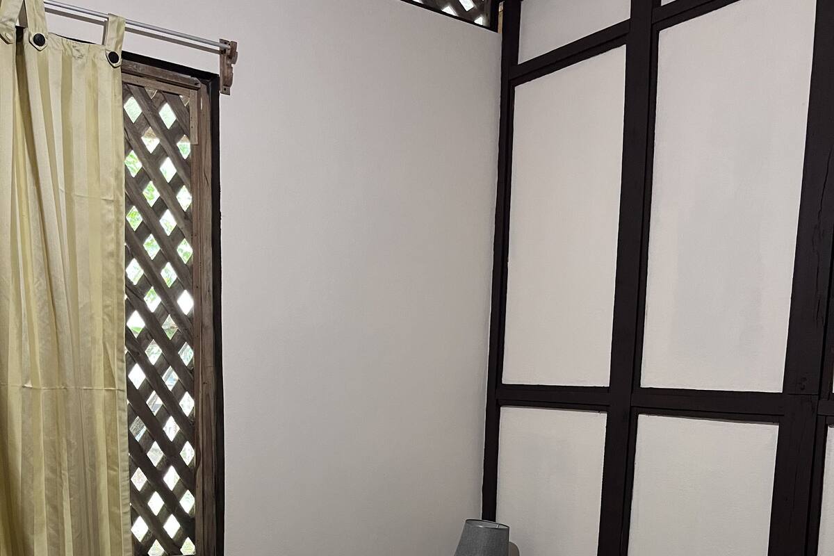 Ein ordentlich bezogenes Bett mit grauer Tagesdecke, einem weißen Kissen und einem gefalteten Handtuch auf der linken Seite. Ein Fenster mit Holzgitterverzierung und halb heruntergezogenem Vorhang sind zu sehen. In der Ecke steht ein kleiner Tisch mit einer Lampe.