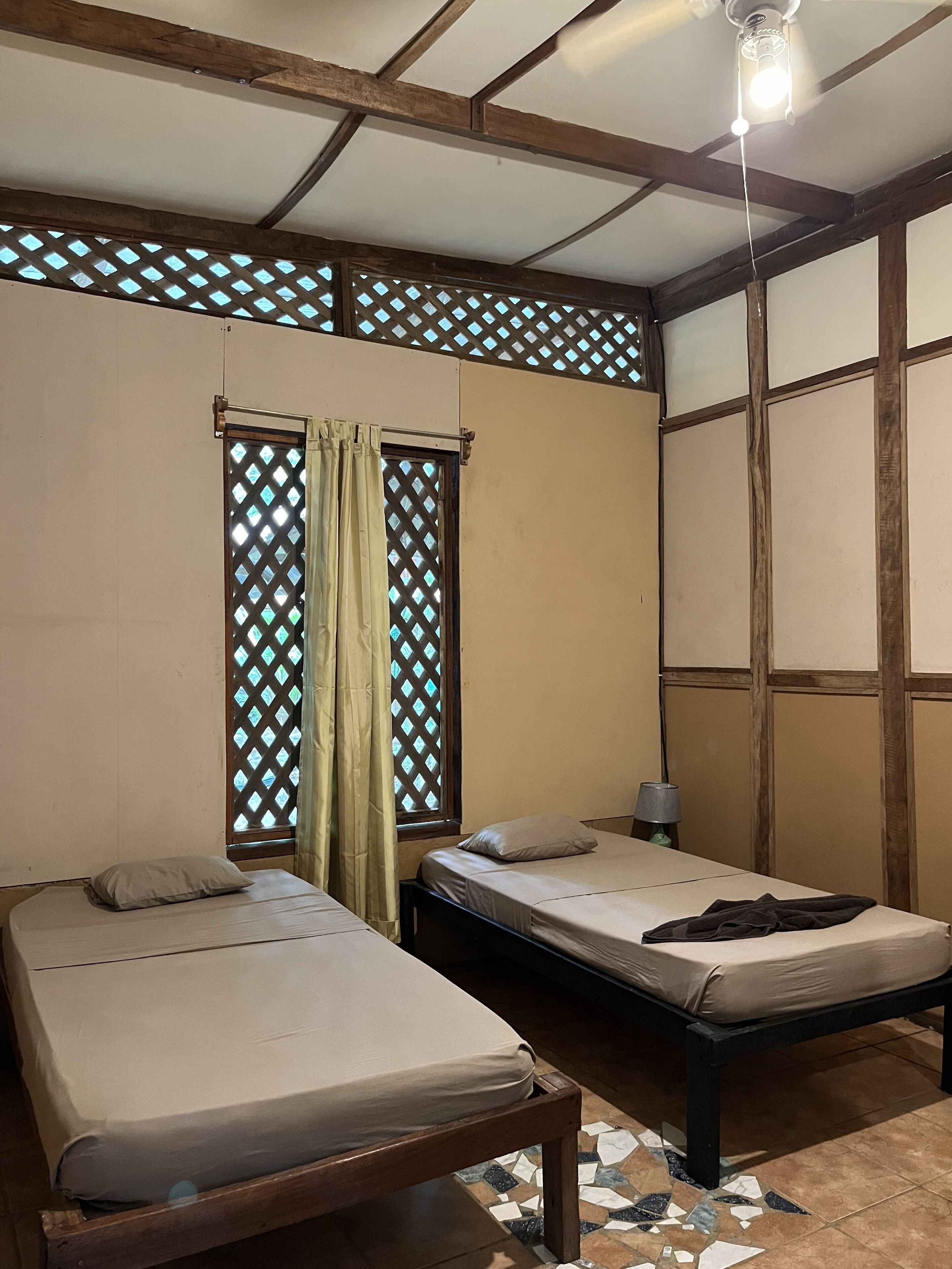 A room with two beds, a window with a lattice design, a ceiling fan, and a tiled floor.