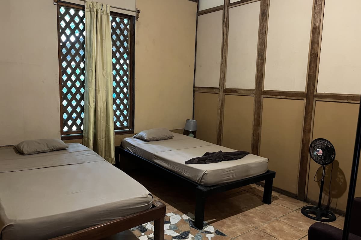Ein Zimmer mit zwei Betten, einem kleinen Tisch, einem Standventilator und einem Fenster mit Gittermuster.