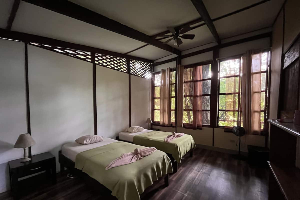 Ein Zimmer mit zwei Betten, einem Deckenventilator und großen Fenstern mit Vorhängen.