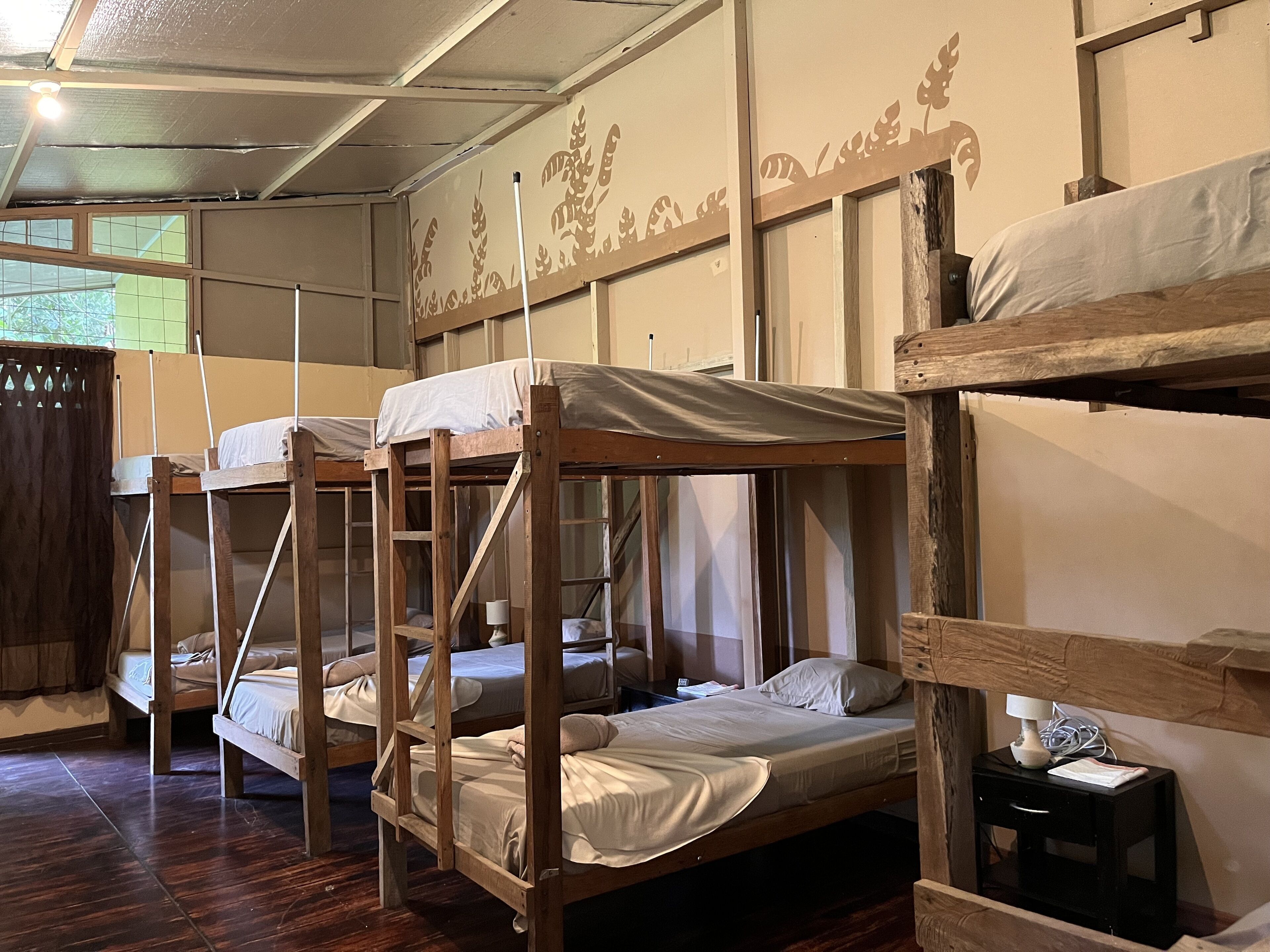 A dormitory room with bunk beds, wooden walls, and a small bedside table with a lamp.