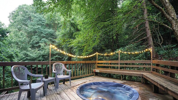 Une terrasse en bois avec un jacuzzi, deux chaises en plastique et des guirlandes lumineuses.