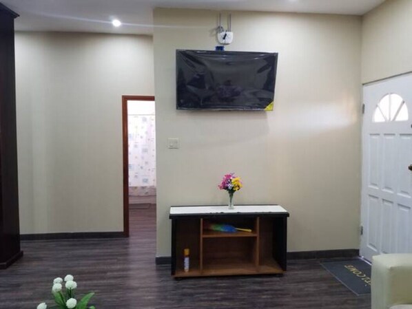 A room with a dark wooden floor, a white door, a small table with a vase of flowers, and a mounted television.