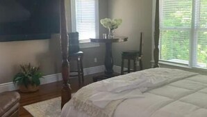A bedroom with a four-poster bed, a ceiling fan, a large window with blinds, a flat-screen TV mounted on the wall, and a small table with a vase of flowers. - A Room Off Broadway, a prime downtown hideaway (Bardstown)