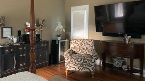 A living room with a four-poster bed, a patterned armchair, a dark wooden dresser, a flat-screen TV, and a window with blinds. - A Room Off Broadway, a prime downtown hideaway (Bardstown)