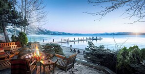 Ein Sitzbereich im Freien am Seeufer mit Feuerstelle, Holzmöbeln und Blick auf einen Steg, der ins Wasser führt.