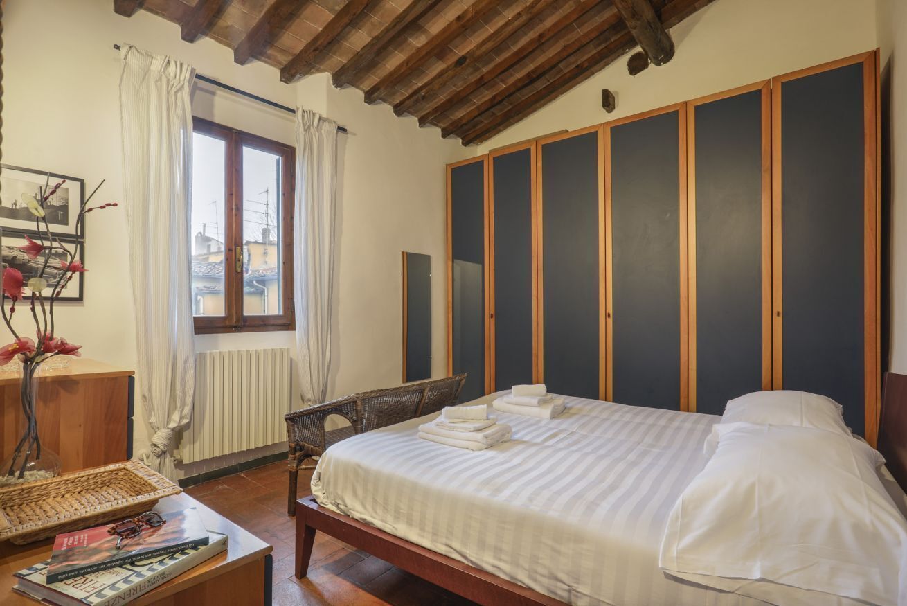 Un dormitorio con cama de madera, ventana con cortinas, una silla, una mesita con un libro y un florero, y una pared con espejo y cuadros enmarcados.
