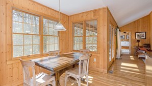 Una cabaña de madera con comedor que incluye una mesa grande y sillas, ventanas amplias y un pasillo que lleva a un dormitorio.
