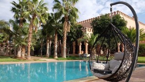 Un espace aménagé au bord de la piscine, avec un fauteuil suspendu, des palmiers et un bâtiment en arrière-plan.