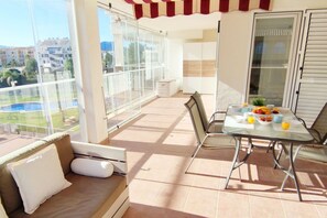 A balcony with a glass door, a cushioned seating area, a table with breakfast items, and a view of buildings and greenery.